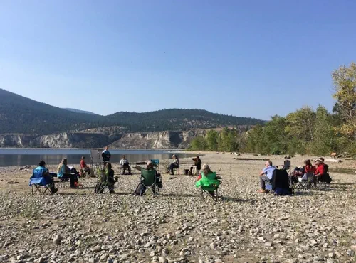 People attending an outdoor session sit in folding chairs on the rocky lakeshore of Gellatly Nut Farm Park, West Kelowna