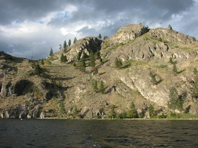 A stretch of Okanagan Lake shoreline featuring a steep, rocky hill dotted with trees and brush.